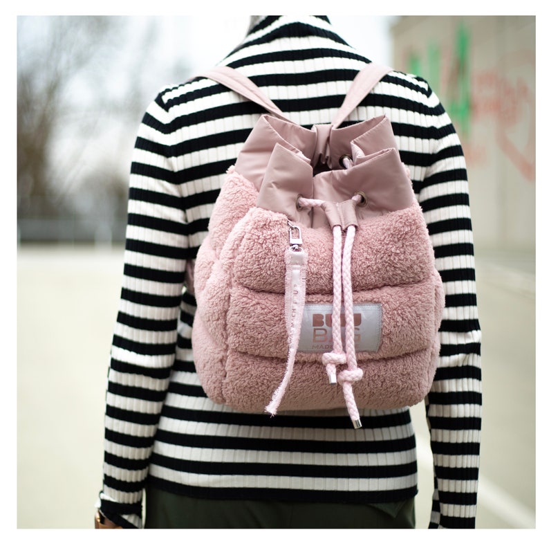 May include: A pink, quilted backpack with a drawstring closure and a fluffy, textured exterior. The backpack has a small, rectangular tag with white text. The straps and drawstring are a lighter shade of pink.