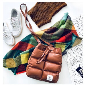 May include: A brown, quilted leather bucket bag with a drawstring closure, a colourful plaid scarf, a pair of white trainers, and a brown knitted neck warmer. The bag has a small tag that reads "CONSUME LESS CREATE MORE."