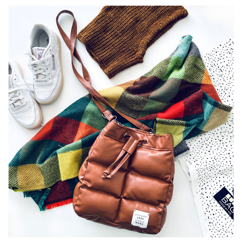 May include: A brown, quilted leather bucket bag with a drawstring closure, a colourful plaid scarf, a pair of white trainers, and a brown knitted neck warmer. The bag has a small tag that reads "CONSUME LESS CREATE MORE."
