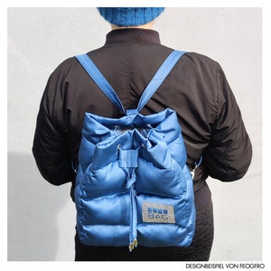 May include: A blue quilted backpack with a drawstring closure and blue straps. The backpack features a silver rectangular patch with the text "LULU BAG". The backpack is worn over a black jacket.