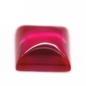 May include: A deep red gemstone with a square shape and a slightly rounded top. The gemstone is cut and polished, and it has a high shine.