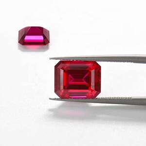 以下が含まれることがあります： ピンセットで保持された深紅色のエメラルドカットの宝石。宝石は鮮やかな赤色で、高い光沢があります。