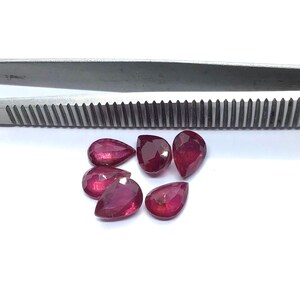 May include: Six pear-shaped ruby gemstones, ranging in size, are arranged on a white surface. The gemstones are a deep red color and have a glossy finish.