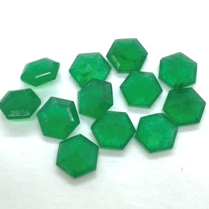 Puede incluir: Un grupo de 11 cabujones de piedra preciosa verde facetados, cada uno con forma de hexágono. Las piedras preciosas están dispuestas en un grupo suelto sobre una superficie blanca.