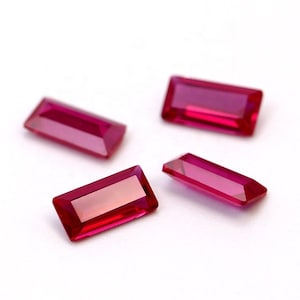 May include: Four rectangular red gemstones with a faceted cut. The gemstones are arranged on a white surface.