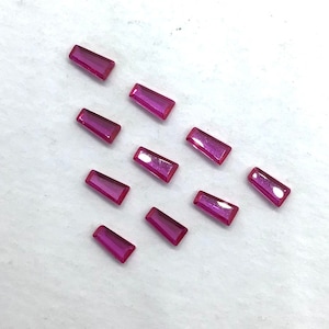 Könnte beinhalten: Zehn rosa rechteckige Edelsteine mit facettiertem Schliff. Die Edelsteine sind diagonal auf einer weißen Oberfläche angeordnet.