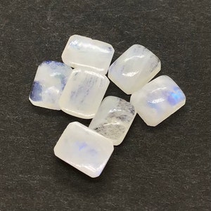 Könnte beinhalten: Sechs regenbogenfarbene weiße Mondstein-Cabochons in rechteckiger Form. Die Steine haben einen blauen Schimmer und sind in einer Gruppe auf einer schwarzen Oberfläche angeordnet.