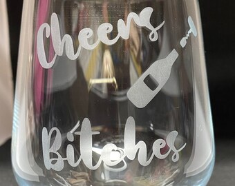 May include: A clear stemless wine glass with the words "Cheers Bitches" etched in a cursive font. A bottle and corkscrew graphic is also etched on the glass. The glass is empty and sits on a dark surface.