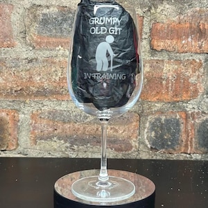 Può includere: Un bicchiere di vino in vetro trasparente con un design in bianco e nero che dice "Grumpy Old Git In Training". Il bicchiere è su una base circolare nera e marrone con il testo "Casa Lucas Glasses".
