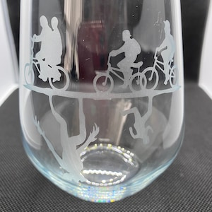 May include: A clear glass wine glass with an etched design of four figures riding bicycles, with the figures upside down below the rim of the glass.