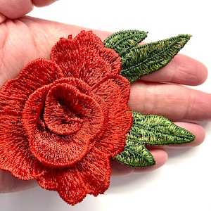 May include: A red embroidered rose with green leaves. The rose is a 3D design with multiple layers of petals.