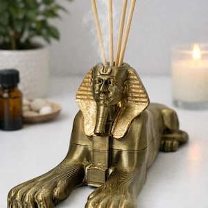 May include: A gold-colored Sphinx-shaped essential oil diffuser with wooden reeds emitting vapor. The diffuser has intricate details and is placed on a white surface. A lit candle and a small plant are in the background.