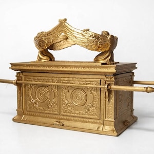 May include: A gold-colored, rectangular chest with ornate carvings and two long poles extending from the sides. Two golden winged figures are perched on top. The chest is likely a decorative item.