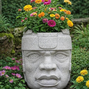 Puede incluir: Una maceta de piedra gris moteada con forma de cabeza humana y una banda ancha. La maceta está llena de un arreglo vibrante de flores de zinnia amarillas, naranjas y rosas, creando un contraste colorido contra la superficie de piedra texturizada.