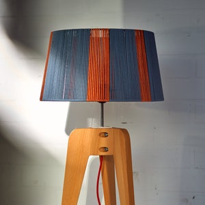 May include: A table lamp with a wooden tripod base and a blue and orange striped lampshade. The lamp has a red cord and a light bulb is on the floor.