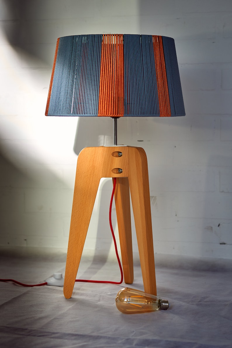 May include: A table lamp with a wooden tripod base and a blue and orange striped lampshade. The lamp has a red cord and a light bulb is on the floor.