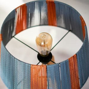May include: A lampshade with a striped pattern of blue and orange yarn. The lampshade is round and has a light bulb inside.