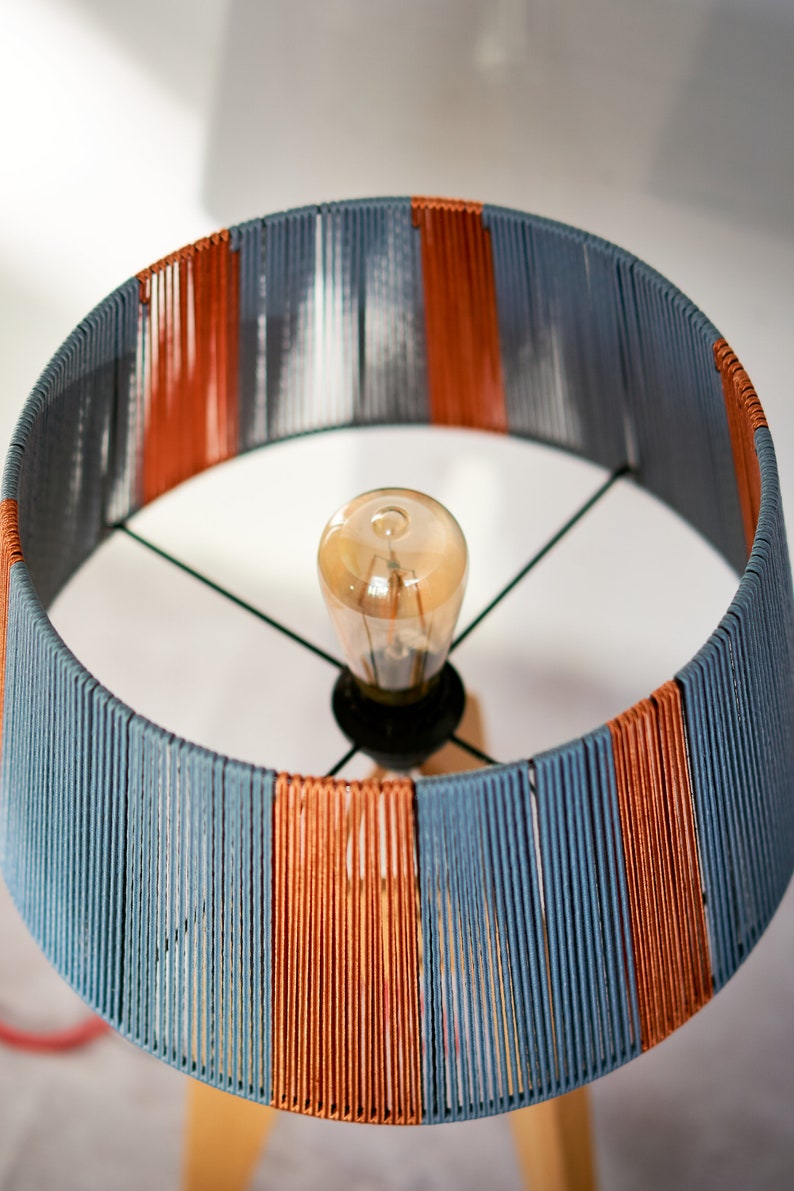 May include: A lampshade with a striped pattern of blue and orange yarn. The lampshade is round and has a light bulb inside.