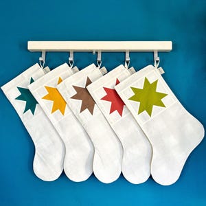 May include: Five white stockings hanging from a light-colored wooden rack with metal hooks. Each stocking features a large, eight-pointed star applique in a different color: dark teal, mustard yellow, brown, red, and lime green.