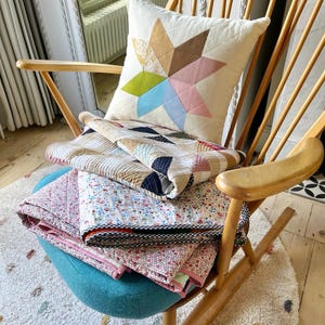Può includere: Una sedia a dondolo in legno con seduta turchese ospita un cuscino a forma di stella e diverse trapunte piegate. Il cuscino presenta un design a stella con vari colori pastello. Le trapunte hanno motivi floreali e geometrici.