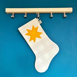 K&ouml;nnte beinhalten: Ein wei&szlig;-hellgrau karierter Weihnachtsstrumpf mit einer gro&szlig;en, leuchtend gelben achtstrahligen Sternapplikation im oberen Bereich. Der Strumpf verf&uuml;gt &uuml;ber ein kleines Stoffetikett mit der Aufschrift LOOM am oberen Rand.