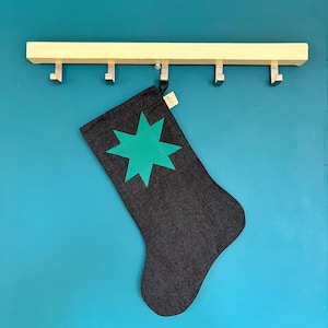 May include: A dark grey fabric stocking featuring a bright teal eight-pointed star appliqu&eacute; on the front. The stocking includes a small beige fabric tag with the text LOOM printed in black, hanging from a light-coloured wooden wall-mounted hook rack.