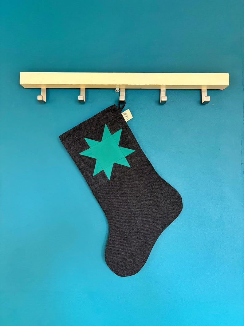 May include: A dark grey fabric stocking featuring a bright teal eight-pointed star appliqu&eacute; on the front. The stocking includes a small beige fabric tag with the text LOOM printed in black, hanging from a light-coloured wooden wall-mounted hook rack.