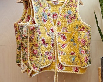 May include: A yellow quilted waistcoat with a floral print and white trim. The waistcoat has a V-neck and ties at the waist.