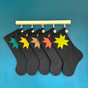 Op de afbeelding: Vijf donkergrijze kerstsokken hangen aan een lichthouten rek. Elke sok is voorzien van een achtpuntige sterapplicatie in een andere kleur: groenblauw, geel, beige, oranje en felgeel. Elke sok heeft een klein wit stoffen label.