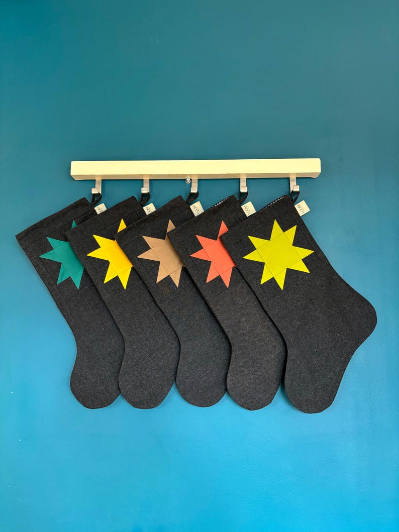 May include: Five dark grey stockings hanging from a light wood rack. Each stocking features a single eight-pointed star appliqu&eacute; in a different colour: teal, yellow, tan, orange, and bright yellow. Each stocking has a small white fabric tag.