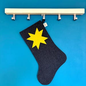 May include: A dark grey fabric stocking featuring a bright yellow eight-pointed star appliqu&eacute; on the front. The stocking includes a small white fabric tag with the text LOOM printed in black, hanging from a light-coloured wooden wall hook rack.