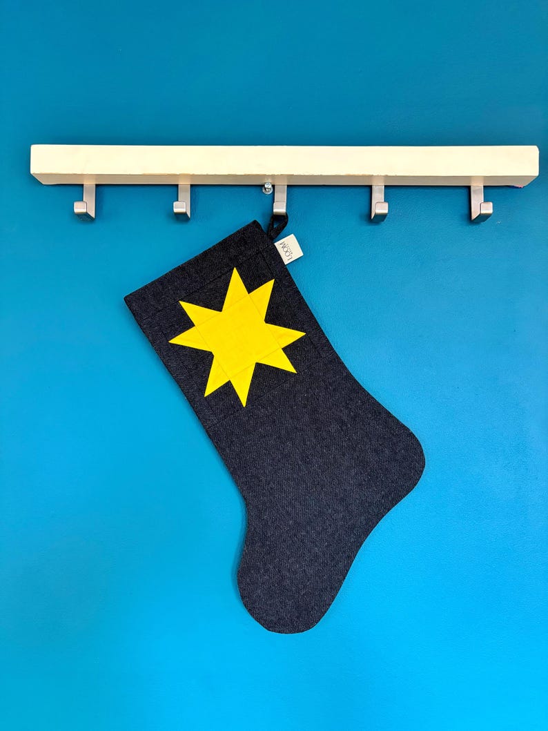May include: A dark grey fabric stocking featuring a bright yellow eight-pointed star appliqu&eacute; on the front. The stocking includes a small white fabric tag with the text LOOM printed in black, hanging from a light-coloured wooden wall hook rack.