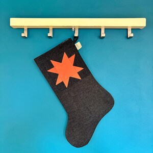 May include: A dark grey fabric Christmas stocking featuring a bright orange eight-pointed star appliqu&eacute; on the upper section. A small white tag with the text "LOOM" is attached to the top edge of the stocking, which hangs from a hook.