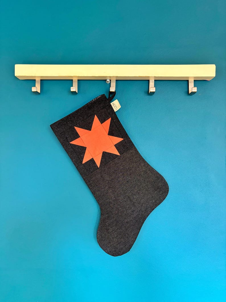 May include: A dark grey fabric Christmas stocking featuring a bright orange eight-pointed star appliqu&eacute; on the upper section. A small white tag with the text "LOOM" is attached to the top edge of the stocking, which hangs from a hook.
