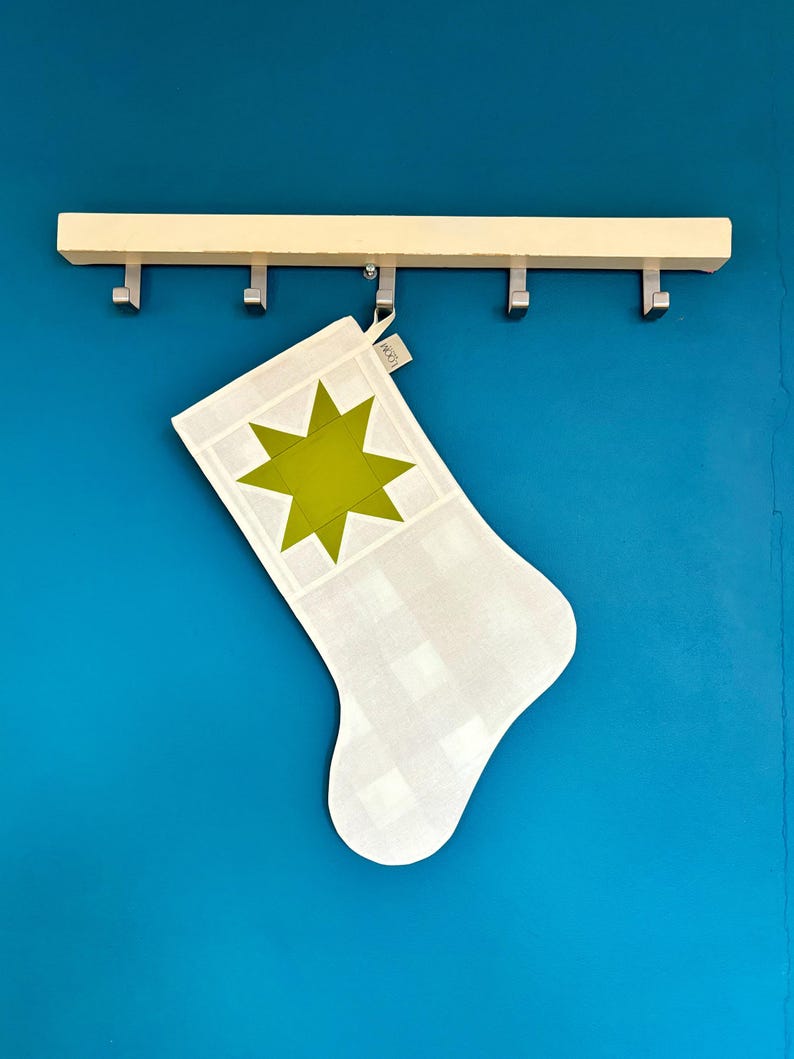 K&ouml;nnte beinhalten: Ein wei&szlig;-hellgrau karierter Weihnachtsstrumpf mit einem gro&szlig;en, einfarbigen, lindgr&uuml;nen achtstrahligen Stern in der Mitte des Umschlags. Ein kleines wei&szlig;es Stoffetikett mit dem Text LOOM ist an der oberen Schlaufe des Strumpfes befestigt.