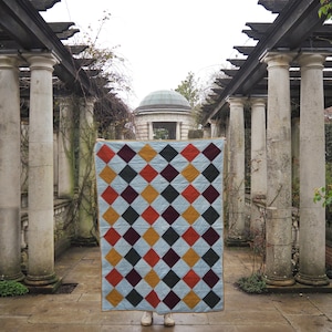 May include: A handmade quilt with a light blue background and a repeating pattern of colorful diamonds in shades of red, orange, yellow, and dark green. The quilt is held up in front of a person in white sneakers.