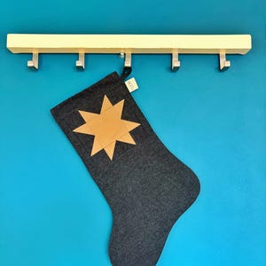 May include: A dark grey fabric Christmas stocking featuring a tan eight-pointed star appliqu&eacute; on the front. A small white tag with the text LOOM is attached to the top edge. The stocking hangs from a light-coloured wooden rack with five metal hooks.