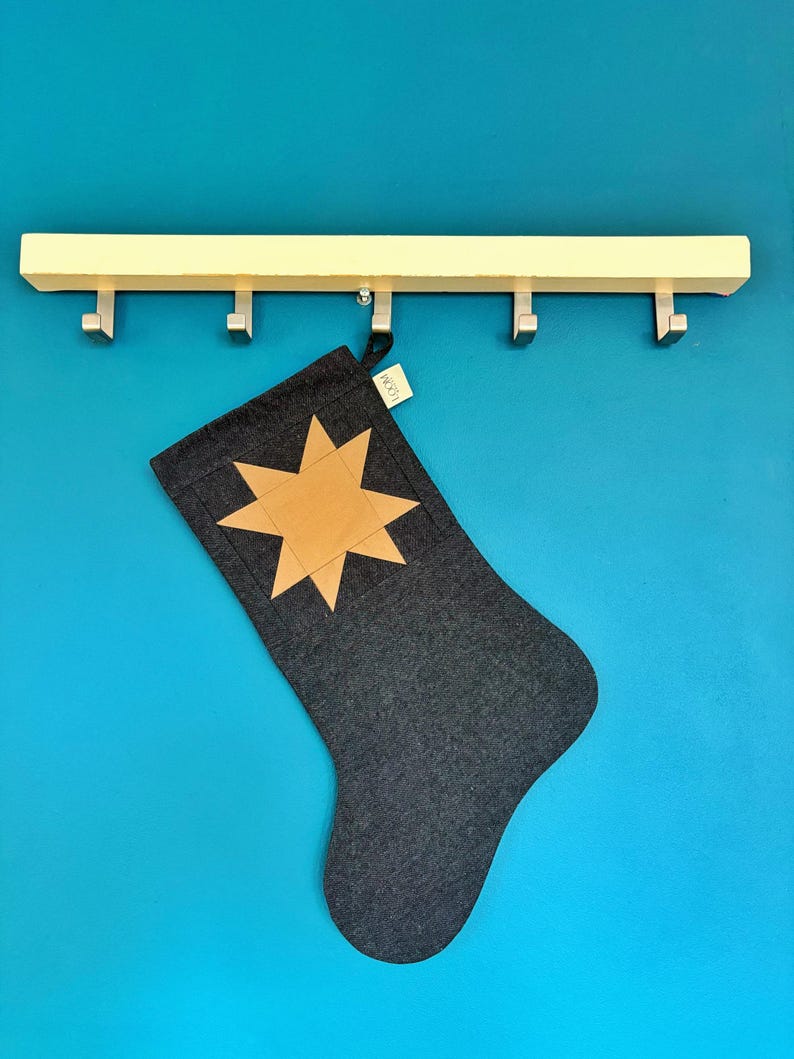 May include: A dark grey fabric Christmas stocking featuring a tan eight-pointed star appliqu&eacute; on the front. A small white tag with the text LOOM is attached to the top edge. The stocking hangs from a light-coloured wooden rack with five metal hooks.