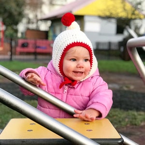 Puede incluir: Un niño pequeño con un gorro de punto blanco y rojo con un pompón en la parte superior, jugando en un parque infantil.