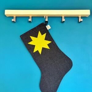 May include: A dark grey fabric stocking featuring a bright yellow eight-pointed star appliqu&eacute; on the front. A small white fabric tag with the text LOOM is attached to the top edge. The stocking hangs from a light-coloured wooden wall hook rack.