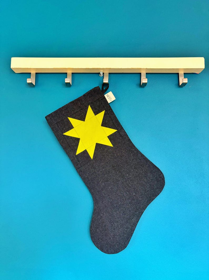 May include: A dark grey fabric stocking featuring a bright yellow eight-pointed star appliqu&eacute; on the front. A small white fabric tag with the text LOOM is attached to the top edge. The stocking hangs from a light-coloured wooden wall hook rack.