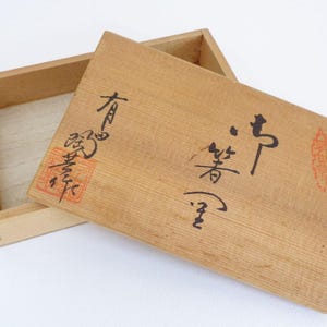 May include: A wooden box with a lid, displaying Japanese calligraphy in black ink. The lid is slightly ajar, showing the box's interior. A red stamp is visible on the lid. The box is made of light-coloured wood.