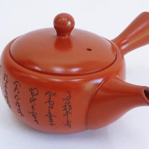 May include: A red clay teapot with a lid and a spout. The teapot has black calligraphy on the side.