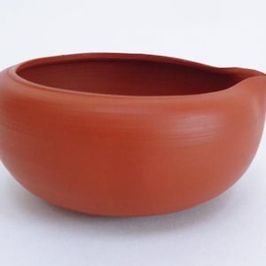 Może przedstawiać: Okrągła ceramiczna miska w kolorze terakoty z dzióbkiem. Miska ma szeroki otwór i gładką, nieszkliwioną powierzchnię. Projekt jest prosty i funkcjonalny, odpowiedni do serwowania lub ekspozycji.