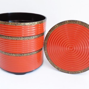 Op de afbeelding: Een Japanse bento-box met drie niveaus, rood gelakt, met een bijpassend deksel. De doos heeft goudkleurige decoratieve randen. Het deksel heeft een concentrisch cirkelontwerp. De binnenkant van de doos is zwart.
