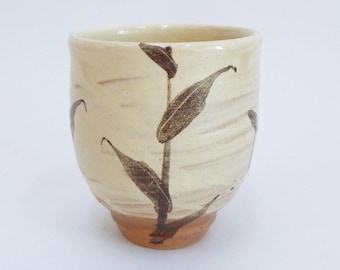 Puede incluir: Una taza de té de cerámica con fondo beige y diseño floral marrón. La taza tiene forma redondeada con borde ligeramente ensanchado y base marrón. El diseño floral presenta hojas largas y delgadas.