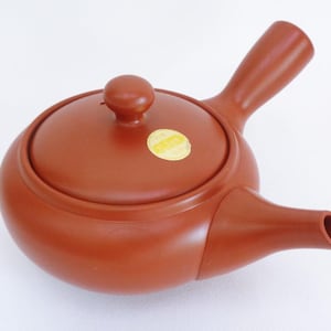 Può includere: Una teiera in ceramica marrone-rossastra con un manico laterale e un beccuccio. La teiera ha un corpo rotondo con un coperchio e un piccolo pomello sulla parte superiore. Un adesivo giallo è sul coperchio. La teiera è progettata per preparare il tè.