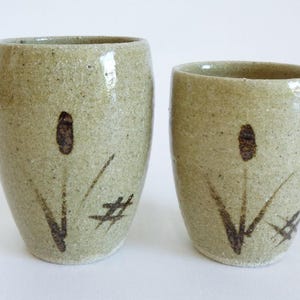 Puede incluir: Dos tazas de cerámica hechas a mano con un esmalte verde claro moteado. Cada taza presenta un diseño de espadaña marrón con líneas finas de color marrón oscuro y un símbolo de hashtag. Las tazas varían ligeramente de tamaño.