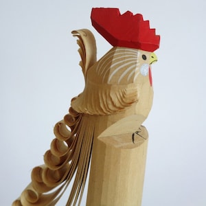 Könnte beinhalten: Eine hölzerne Hahnfigur mit einem roten Kamm und einem detaillierten, gekräuselten Schwanz. Der Hahn ist mit weißen Streifen und einem gelben Schnabel bemalt.