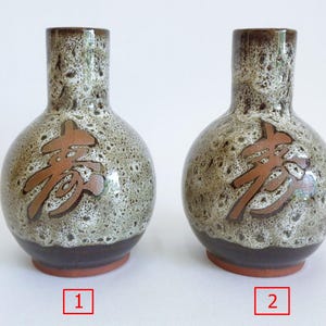 May include: Two ceramic vases with a speckled, earthy-toned glaze. Each vase features a brown, stylized character on the front. The vases have a dark brown base and a reddish-brown bottom. The vases are displayed on a white surface.